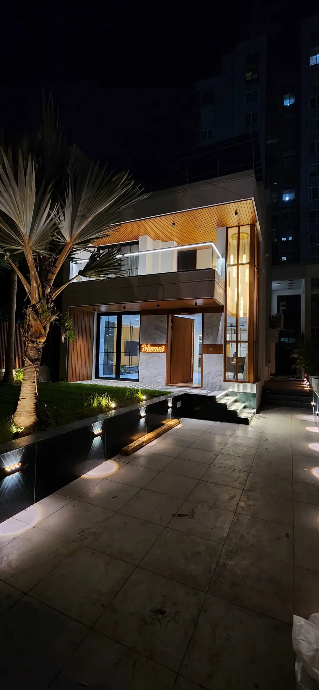 Contemporary bungalow at night with a glazed entrance, tall vertical glazing and a wood-clad balcony, lit along a paved garden path