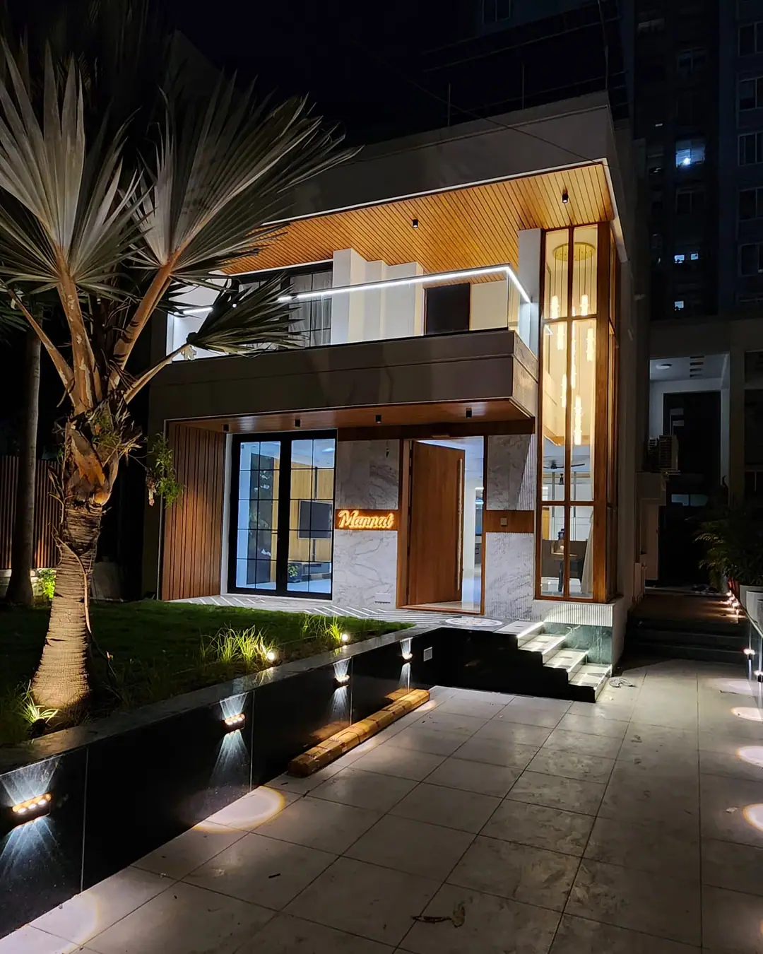 Contemporary bungalow at night with a glazed entrance, tall vertical glazing and a wood-clad balcony, lit along a paved garden path