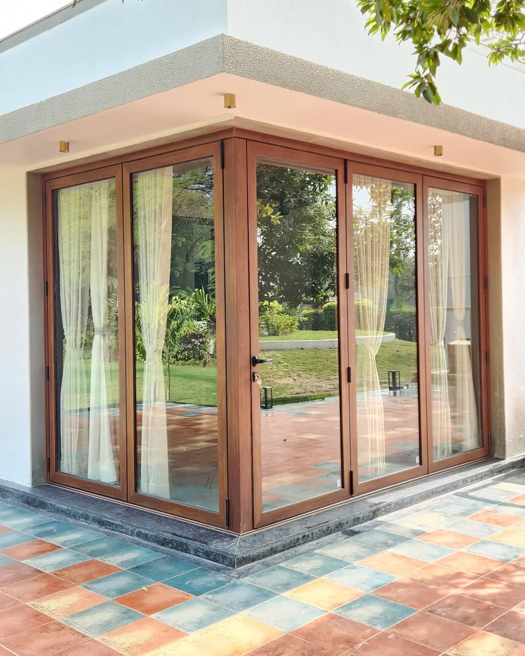 Wood-finish aluminium glazed doors wrapping the corner of a house, opening onto a terracotta-tiled patio and garden
